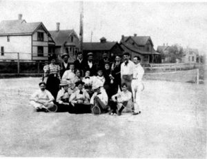 Grayscale long shot of a tennis team in Victorian sportswears posing in an outdoor court. Some carry tennis racquets. People in the front sit on the ground. A line of buildings is visible in the far background.