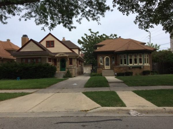 Long shot of two bungalows separated by a driveway. Both feature one-and-a-half-story buildings with an arched entrance door. The buildings have front yards with green lawns.