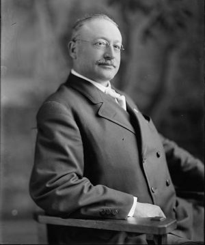 Portrait of Victor Berger in grayscale tone sitting facing slightly to the right in an armchair. He wears round glasses and a curvy collared suit. Both his arms rest on the chair. His eyes look to the right radiating confidence.
