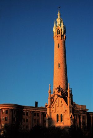 Long shot of North Point Water Tower hit by orange sunshine coming from the left. The tower's base has two arched windows and some buttresses crowned with pinnacles. A dragon sculpture is perched atop the base. The tower's shaft features other round-arched windows. One of the top portion's pointed arched windows is visible. A large building and a blue sky appear behind.