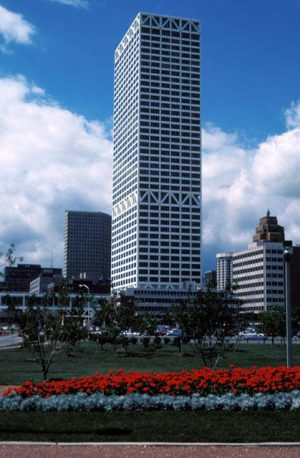 Long shot of the U.S. Bank Center tower soaring above other buildings in the vicinity. The Lakefront Park is in the image's foreground. Several green trees and red-colored landscaping plants grow in the park. The blue sky is above.