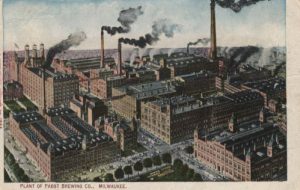 A painted postcard illustrates a bird's eye view of the Pabst Brewing Company plant. The giant building complex has several chimneys that release steam and black smoke. Other buildings and the city's roadways are visible in the surrounding area. Text at the bottom of the postcard reads "Plant of Pabst Brewing Co. Milwaukee."