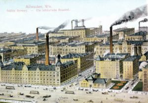 A painted postcard showcases an aerial view of the Schlitz Brewing Company plant and the surrounding area. All buildings face slightly to the left. The factory's chimneys billow black smoke to the right. Horse-drawn carriages and other vehicles traverse the streets around the area in the foreground.