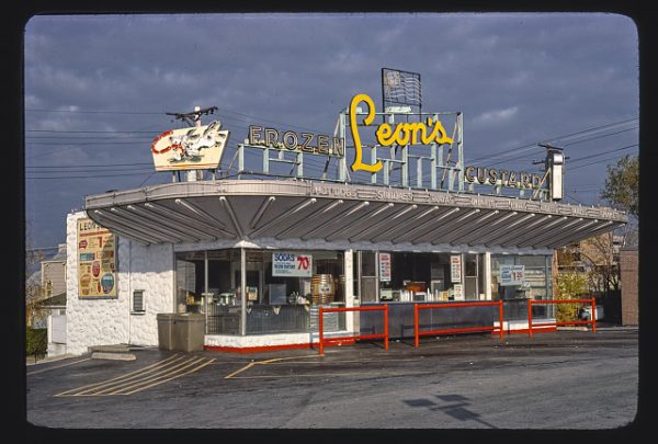 Exterior view of Leon's Frozen Custard stand and drive-in against a cloudy sky. The large yellow store sign is installed on top of the building. Different kinds of equipment placed right next to the store's glass wall are visible from the outside.