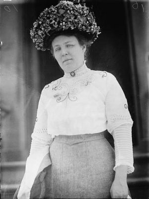 Grayscale medium shot of Meta Schlichting Berger from the thighs up standing in a long sleeved blouse with high rigid collars. She wears a large hat adorned with floral pieces.