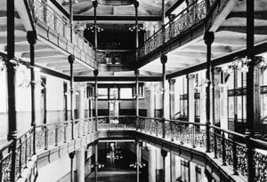 Grayscale long shot of the City Hall interior displaying a portion of its extensive eight-story open atrium. This image shows the top five stories and the ceiling. Each floor is enclosed by balustrades with detailed lace ironwork.