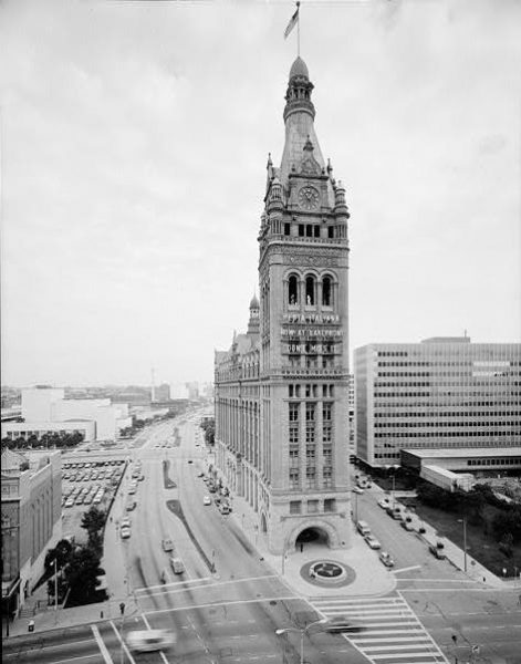 Panoramic view of the City Hall facade from the South side in grayscale tone. This massive building ornamented with a massive bell tower appears prominently among other buildings in the area. Far below, cars drive and are parked on the side of the surrounding streets.