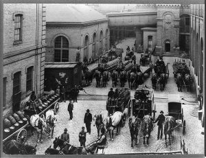 Grayscale high-angle shot of horse-drawn carriages carrying kegs of beer on a brick-paved street next to Schlitz Brewery buildings. Some people stand on the road, some sit on the carriages.