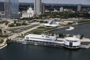 Bird's eye view of Discovery World's white-colored structures standing on a peninsula protruding into Lake Michigan. A group of boats is docked near the peninsula. The Milwaukee Art Museum's building and the city's eastern skyline appear in the background.