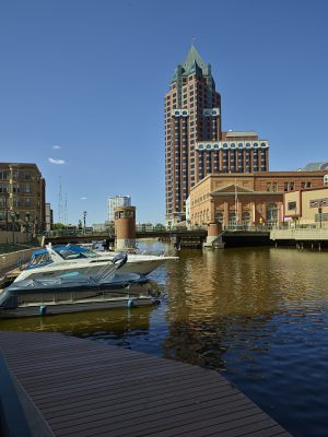 Long shot of boats docked on the left in the Milwaukee River. The water body flows from back to front. A bridge and downtown buildings are visible in the background. A clear blue sky spans above.
