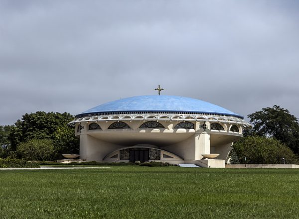 Long shot of the Annunciation Greek Orthodox Church facade with a blue domed roof and a cross on top on a cloudy day. The building is set between groups of green trees. A manicured green lawn is in the foreground.