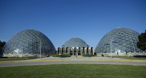 A long shot of three glass domes glistening in daylight, absorbing the blue color from the sky above. The center dome is located a bit far behind. In front of it, flanked by the left and right domes, is a series of nine arched structures, each having a set of windows. Only three structures in the center feature an entrance. Landscaping plants and benches are also situated between the left and right domes. A road stretches from left to right in the foreground. A green lawn appears at the image's bottom portion.