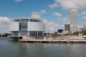 Photograph of Discovery World showcases its iconic circular aquarium on the left. Adjacent to it is the seating area of an outdoor event venue. This arena sits on Milwaukee's lake shore. The US Bank Building and the blue sky are in the background.