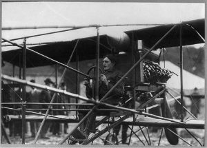 Wide shot of Charles K. Hamilton showing his posture sitting on his vintage airplane called the Hamiltonian parked on flat ground. He wears a long-sleeved jacket and boots. His hands are attached to the plane's circular steering wheel while one of his feet steps on a part of the aircraft. On top of his head and behind his back is the plane's engine. Several blurry figures stand at the rear of the aircraft.