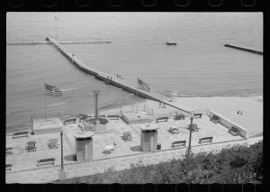 High-angle shot of a beach taken from higher ground. The calm water surface fills up the upper portion of this image. A breakwater structure with a walkway appears in the middle. A group of picnic tables and benches are placed on a rooftop patio of a building erected on the shore. Two flapping American flag stand on it. Some people sit on the sand below.