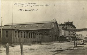 Sepia-colored long shot of a wooden building facing to the right. The building's elongated wings are visible. A large banner hanging over the street in front of the building reads "Soldiers of the Union, Welcome Home." The street spans from the right background to the left foreground. Text at the top right reads "West Side of Broadway, Extending Across Huron St. 1865, The Great Fair."