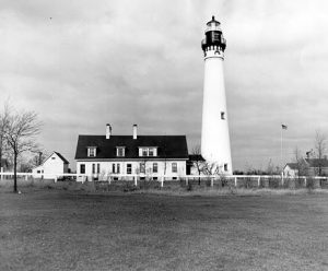 Long shot of Wind Point Lighthouse in grayscale. In the far back is a one-and-a-half-story building with dormer windows, which used to be the keeper's house. A small structure with a gable roof stands on the left of the house. A covered walking path connects the dwelling with the sturdy lighthouse tower on the right. The tower has a lightning rod, beacons, windows, and an observation deck. A long fence span from left to right separates the lighthouse complex from the empty ground in the foreground.