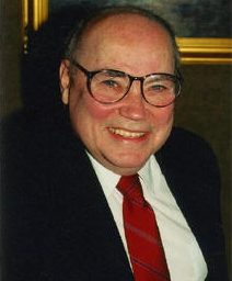 Headshot of Eldon Murray smiling in formal attire and glasses, making eye contact with the camera lens. Murray wears a black suit, a red tie, and a white dress shirt.