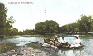 A painted postcard depicts three people on a rowboat on the Milwaukee River. A small portion of land appears in the left to center foreground. The river spans from the left back to the right foreground. Lush green trees soar in the background.
