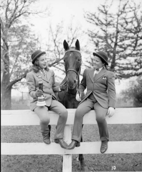 A grayscale image of two girls sitting on the fence in an outdoor space with a horse's appearing between them. The girls wear formal suits, ties and round hats. The one on the left carries a small trophy with her right hand and touches the animal's head with the other hand. The other girl smiles while holding the horse's reins.