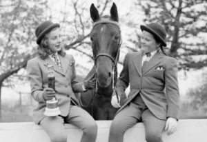A grayscale image of two girls sitting on the fence in an outdoor space with a horse's appearing between them. The girls wear formal suits, ties and round hats. The one on the left carries a small trophy with her right hand and touches the animal's head with the other hand. The other girl smiles while holding the horse's reins.