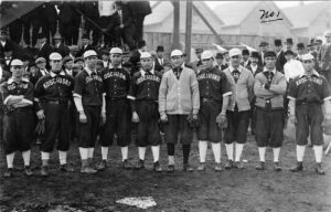 Ten men stand in a row in baseball uniforms that feature the word "Kosciuszko." Some wear jackets and baseball gloves. Behind them are a bunch of men in suits posing for this photograph.