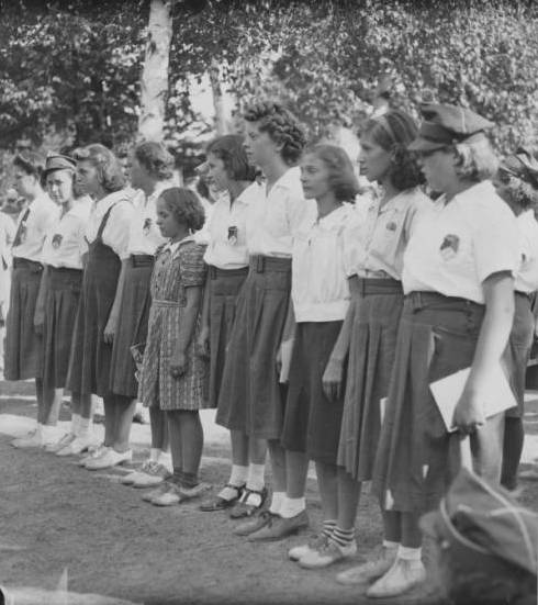 Side view of ten female scouts standing side by side in an open space in grayscale. Most of them are in skirts and short-sleeved shirts. One person, who is in a different dress, stands in the middle. Some people and trees are visible in the background.