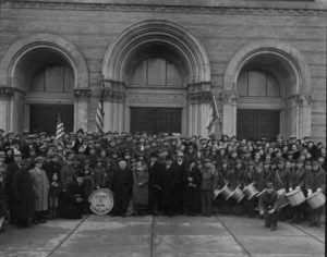 Group photo of a crowd of Polish National Alliance Scouts in grayscale. They pose in front of a building with three large arched doors. Several people on the right side of the front row stand while carrying drums. Two American flags are visible.