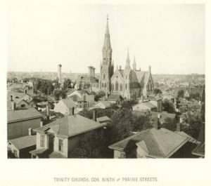 Elevated view of the Trinity Evangelical Lutheran Church building soaring in the distance. Other buildings are visible in the foreground and around the church in the background. Beneath this sepia-colored photograph is a text that reads "Trinity Church, Cor. Ninth, and Prairie Streets."