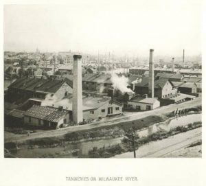 An elevated view of tanneries near the bank of the Milwaukee River in sepia. Billowing steam appears from the tanneries' roof. The water body spans the foreground. Several tall chimneys stand among the buildings.