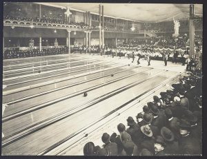 Long shot of the interior of a bowling alley full of spectators watching the bowlers in action. The long bowling lane looks dominant in this black and white photograph, filling the frame from the left side until almost the entire right side of the image. Some players stand still, watching the balls they have just thrown rolling down the bowling lanes. A replica of the Statue of Liberty is visible in the background of the spectator stands.