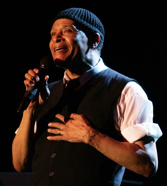 Medium shot of Al Jarreau from the waist up facing left in a light-colored button-down shirt and dark vest; the shirt sleeves are rolled to above his elbows. Jarreau holds a microphone with his right hand and touches his chest with left hand as he performs against a dark background.