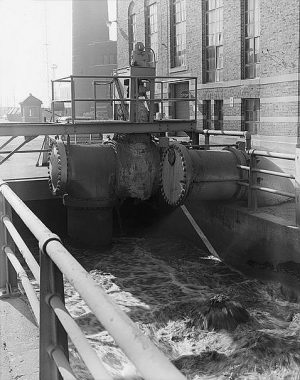 Grayscale photograph of a pump and mixing channel filled with turbulent water below. A railing enclosing the channel stretches on the left-to-center foreground. A portion of a building's exterior wall is in the background.