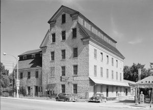 Exterior view of Cedarburg Mill facing left next to a small filling station. This grayscale image shows the five-story building that has one wing on the left. Two cars are parked by the building. The structure stands out in its surrounding area.