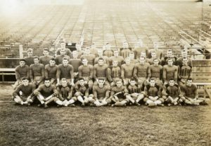 Sepia-colored photograph of the 1936 Marquette University football team in the Cotton Bowl stadium. Four rows of the men in their uniforms pose with backs to the empty bleachers.