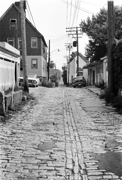 Image of a long alley with bumpy paving slabs. Houses along with power poles appear on the left and right sides of the road. A light-colored car is parked by the left edge of the alley, and a darker-colored one is placed on the opposite side.