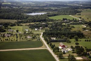 Aerial shot of the Town of Erin displaying a wide rural farm area and a few dwellings among the green grounds. A lake surrounded by groves of trees is visible in the background.