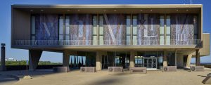 Wide shot of the Milwaukee County War Memorial Center building against the clear blue sky. This image showcases the building's entrance on the ground level and five large mosaic panels on the upper floor enclosed with balustrades. Each mosaic panel displays a combination of Roman numerals in large size. Six stone monument signs stand in front of the entrance. From left to right, the signs are titled "U.S. Army," "U.S. Marine Corps," "U.S. Navy," "Milwaukee County War Memorial," "U.S. Air Force," and "U.S. Coast Guard."