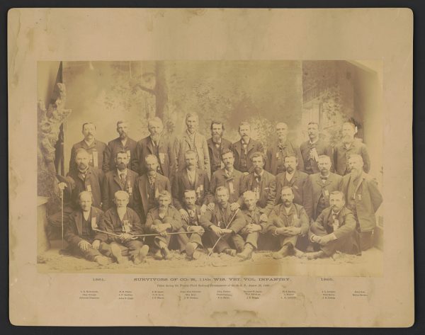 Sepia-colored group photo of survivors of Co. H, 11th Wisconsin Volunteer Infantry posing in three rows. Men in the front row are seated on the floor. Men in the second row are kneeling and those in the back standing. They all make eye contact with the camera lens. Text beneath the photo includes the names of all the veterans.