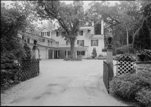 Grayscale long shot of Ten Chimneys residential building and its surroundings. The front gate in the image's foreground is open, showcasing the driveway towards the house in the background. Tall trees grow near the dwelling. Landscaping plants grow by the gate and the house.