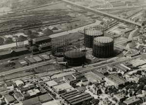 Aerial shot of the Menomonee Valley in sepia. The Wisconsin Gas Company's coal gas processing and storage facility sits at the image's center. A water body is in the background. Several long roadways are located near the facility. Some residential places appear in the foreground.