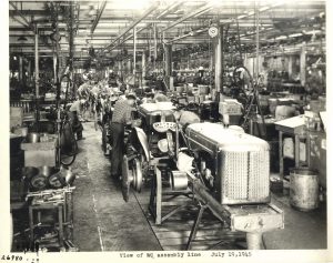 Sepia-colored photo of Allis-Chalmers factory's interior displaying the employees assembling WC model tractors along the image's center. A row of other types of equipment appears on the left and right sides. Streel structure is visible on the ceiling. Texts beneath the image, at the bottom center, read, "View of WC assembly line" and "July 19, 1945."