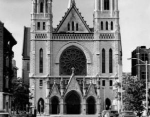 Grayscale long shot of the north facade of Gesu Church. The central building is flanked by two iconic towers topped by crosses. The taller tower, on the right, has a clock. This image shows the main entrance with three arched openings atop four columns. A huge rose window is installed above the entrance.