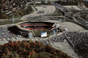 Bird's eye view of Milwaukee County Stadium with a green field on its ground surrounded by empty, multilevel grandstands. Rows of cars are parked in large parking lots outside the stadium. A portion of a green landscape appears in the foreground. Crisscrossing highways are in the far right background.