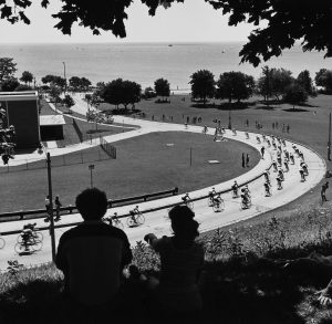 High-angle shot of a bicycle race from the viewpoint of the spectators who sit on the hill. This grayscale image shows two people sitting on the grass under a tree shade, watching the racers heading down the hill on a winding road. The vast expanse of Lake Michigan appears in the distance, in the upper portion of this picture.