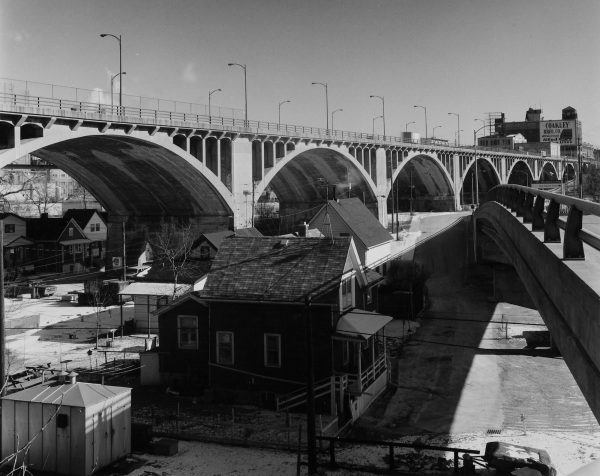 The old Wisconsin Avenue viaduct stretches from the left to right background in grayscale tone. A tall building with a "Coakley Bros. Co" ad appears in the far right background. The guard rails of an overpass are visible on the right. Several buildings below the viaduct appear in the far left back. Several others can be seen in the foreground, in the area between the viaduct and the overpass.