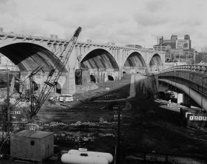 Grayscale long shot of Wisconsin Avenue viaduct during reconstruction. The structure spans from the left to right background. A tall building with a "Coakley Bros. Co" ad appears in the far right background. The guard rails of an overpass are visible in the right foreground. Heavy equipment and a truck are in the area between the viaduct and the overpass. There are almost no buildings in this area.
