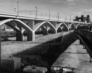 Grayscale long shot of the Wisconsin Avenue viaduct spanning from the left to right background. A tall building with a "Coakley Bros. Co" ad appears in the far right background. The guard rails of an overpass are visible in the right foreground. Grasses and a road can be seen in the area between the viaduct and the overpass.