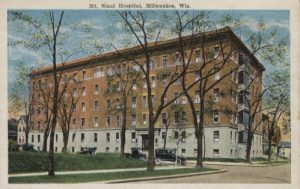 A painted postcard illustrates the exterior view of Mount Sinai Hospital in the distance. The rectangular multi-story building sits on a street corner. The ground floor has a cream-colored exterior wall, while the floors above feature a brown color. Several vintage cars line the street next to the hospital. Tall trees with sparse foliage grow on the lawn around the building. A green space appears in the left foreground. A portion of an empty street is on the right front. The blue sky is depicted above.