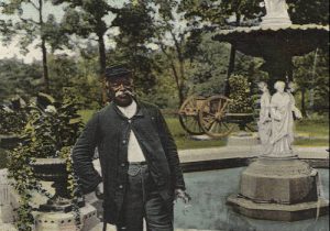 Postcard depicting an African-American man standing with a walking stick posing next to a fountain. He faces directly to the camera lens in a hat, jacket, and trousers. The fountain features two layers of sculptures of women in flowing gowns. Green lawn and trees are in the background.